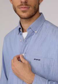 Chemise button-down bleu clair en tissu doux, dotée d'une poche poitrine avec trois silhouettes d'animaux brodées et d'un col à boutons.