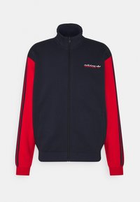 Chaqueta de cremallera en azul marino y rojo con mangas largas, que presenta tres rayas negras en cada brazo y un logo bordado en el pecho.