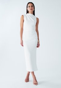 Robe midi blanche sans manches avec un design drapé, une texture lisse et une silhouette ajustée. Portée avec des sandales à talons hauts nude à bretelles.