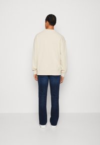 Man with braided hair wearing a cream sweatshirt, dark blue jeans, and white shoes, standing facing a plain light background.