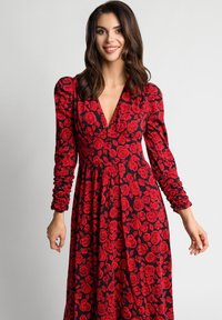 Kobieta z brązowymi włosami, ubrana w czarną sukienkę z długim rękawem w czerwone róże, uśmiechająca się i stojąca na tle jasnej, jednolitej powierzchni.