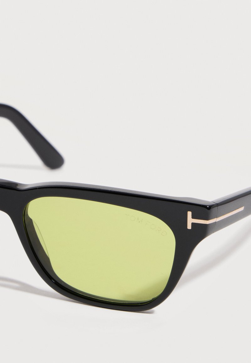 Lunettes de soleil rectangulaires noires avec des verres teintés jaune-vert et un logo "T" doré sur les branches, sur fond blanc.