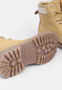 Paire de bottes de randonnée couleur bronzée avec semelles en caoutchouc épais et texturé, présentant des crampons profonds et des colliers de cheville rembourrés.