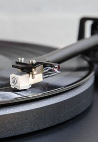 Black turntable close-up featuring a silver and white cartridge, a stylus, and colored wiring. Round platter with a smooth finish.