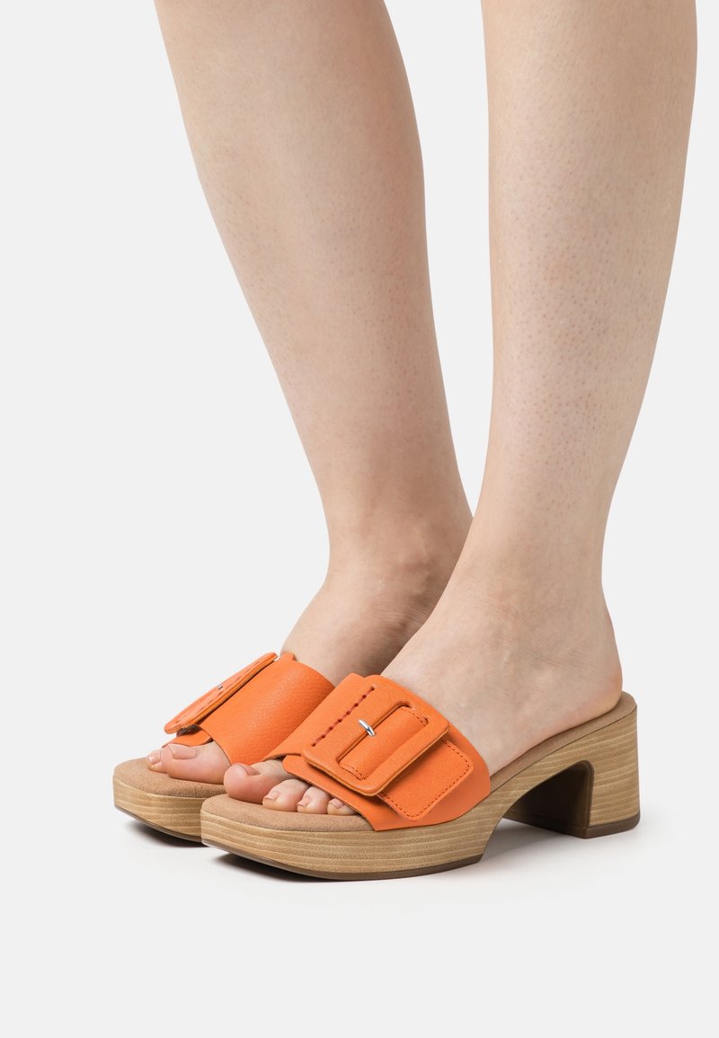 Pieds chaussés de sandales ouvertes en cuir orange avec de larges brides à boucle et des talons blocs en bois épais sur fond blanc.