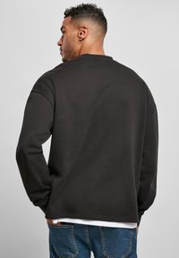 Hombre con cabello corto y rizado, y arete de aro, que lleva un sudadera negra sobre una camisa blanca y jeans azules, de espaldas con las manos en los bolsillos.