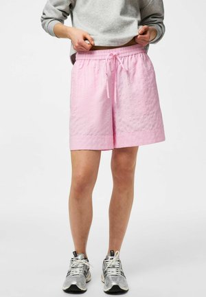 Osoba ubrana w jasnoróżowe, teksturowane szorty ze ściągaczem, szarą bluzę i srebrne sportowe buty, stojąca na jednolitym białym tle.