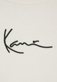 Vit tyg med svart broderad signaturdesign, som har mjuka linjer och varierande tjocklekar i den kursiva skriften.