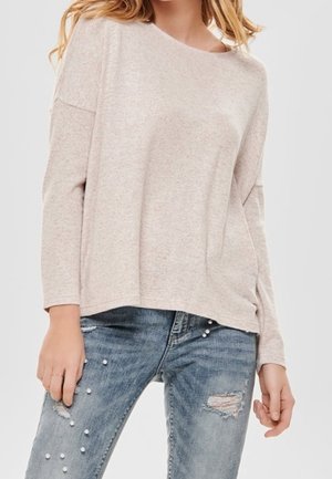 Femme portant un haut ample à manches longues de couleur beige clair, associé à un jean bleu déchiré orné de petites perles.