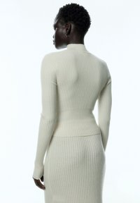 Rippstrick-Cremepullover mit hohem Ausschnitt und figurbetontem Schnitt. Lange Ärmel und eine glatte Textur. Getragen mit einem passenden Ripprock.