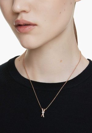 Collier en or rose au design entrelacé, avec de petits accents. Porté sur un pull noir, mettant en valeur la texture délicate de la chaîne.