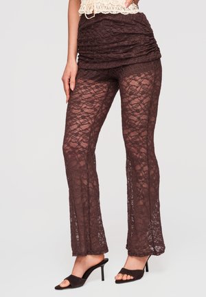 Femme portant un pantalon en dentelle marron transparent avec un short froncé par-dessus et des mules noires à talons ouvertes, debout devant un fond neutre.