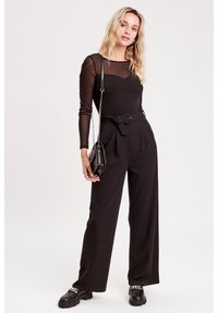 Haut à manches longues en maille noire avec texture à pois, associé à un pantalon noir taille haute. Un petit sac à main noir et des chaussures massives complètent le look.