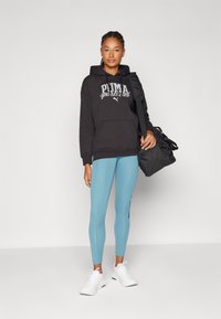 Felpa nera con stampa "PUMA sports club", leggings azzurri chiari e sneakers bianche. Borsa da palestra nera con tracolla.