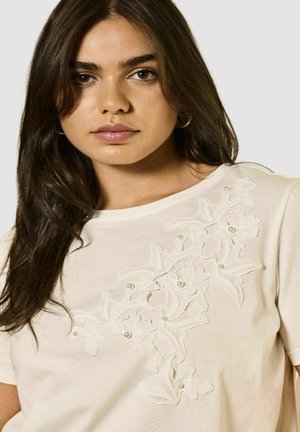 T-shirt crème avec broderies florales, accents texturés et petits détails en perles, col rond et manches courtes.