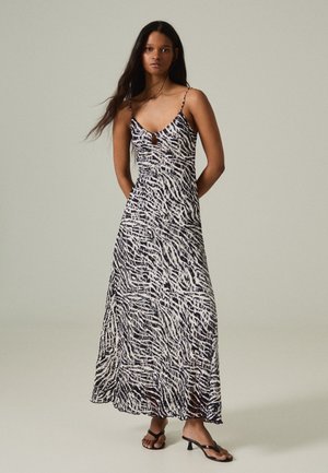 Femme portant une longue robe imprimé zèbre noir et blanc avec de fines bretelles et des sandales noires à talons plats, debout devant un fond uni.