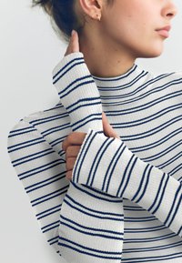 Pulover s polo ovratnikom s črtami v beli in mornarsko modri barvi, z rebrasto teksturo in podaljšanimi rokavi z luknjami za palce, ki izboljšujejo udobje in prileganje.