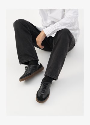 Svarte joggesko med en glatt og teksturert overflate, med gummisåle og mørke snører. Kombinert med svarte jeans og en hvit stripet skjorte.