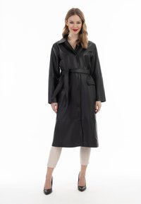 Trench coat in pelle sintetica nera con maniche lunghe, chiusura a bottoni, cintura legata in vita e tasche laterali. Modello che indossa pantaloni crema e tacchi.