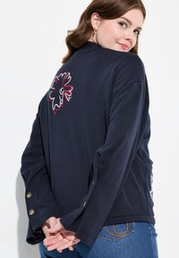Marinblå långärmad topp med blommig brodering på baksidan, knäppta manschetter och en avslappnad passform, kombinerad med denimjeans.