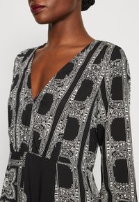 Robe noire avec un décolleté en V profond, ornée de motifs blancs complexes et de rayures verticales. Manches longues et texture de tissu douce et lisse.