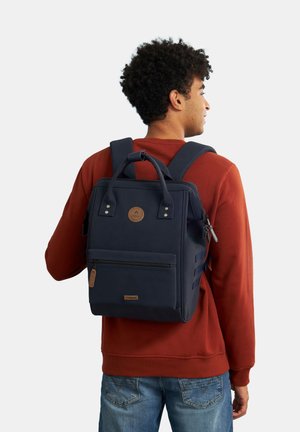 Sac à dos bleu marine avec poignée supérieure, poche zippée à l'avant et sangles latérales. Fabriqué en tissu durable avec une finition texturée et un détail de logo.