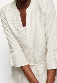 Robe beige clair avec de subtiles rayures blanches, présentant un col en V, des manches évasées et des accents volantés à la taille et aux ourlets des manches.