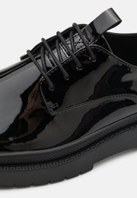 Zapato de charol negro con punta redonda, que presenta cordones redondos negros, una superficie lisa y una suela texturizada.