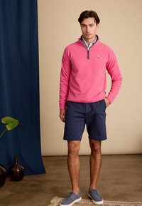 Lichtroze pullover met een ritskraag, gecombineerd met marineblauwe shorts. Model in grijsgroene schoenen, staande op een gestructureerd oppervlak.