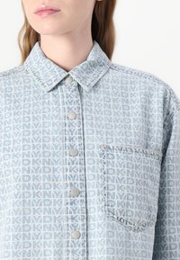 Light blue denim shirt featuring a geometric pattern, button closure, and single chest pocket. Smooth texture with a collared design.