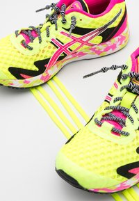 Des chaussures de sport néon jaunes avec des accents noirs et roses, en matériau en maille, semelle à motifs et lacets distinctifs rayés noirs et blancs.