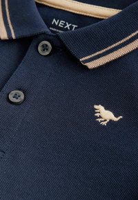 Jūrų laivyno spalvos polo marškiniai su smėlio spalvos juostele ant apykaklės, dviem juodais mygtukais ir mažai smėlio spalvos siuvinėtu dinozauru ant kairiojo krūtinės.