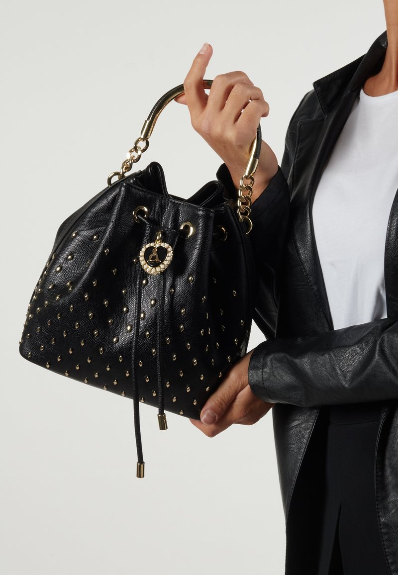 Mustakankainen nahkainen ämpärilaukku, jossa on kultaisia niittejä, nyörisuljenta ja ketjuhandle, jossa on koristeellinen pyöreä yksityiskohta edessä.