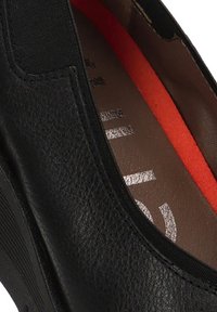 Zapato negro tipo mocasín que presenta un upper de cuero texturizado, paneles elásticos en los laterales y una plantilla roja contrastante con un logotipo impreso.