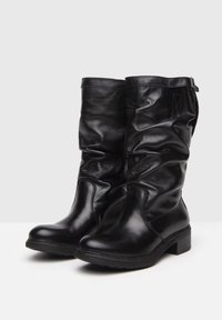 Mustat nahkaiset polvimittaiset saappaat, joissa on laskostettu muotoilu, pyöristetty kärki ja lähellä oleva korko. Huomattavaa ompelua ja pieni koriste ylhäällä.