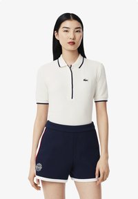 Vit polo skjorta med korta ärmar, marinblå accent och Lacoste-logotyp. Matchas med marinblå shorts med vita och röda sidostripes och logotyrläggning.