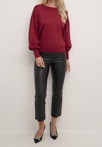 Pull rouge drapé avec un col rond et des manches longues, associé à un pantalon noir en simili cuir coupé et des talons noirs à bout pointu avec des accents.