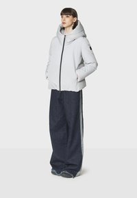 Giacca puffer grigio chiaro con cappuccio oversize e zip nera, abbinata a pantaloni wide-leg in denim scuro con righe laterali grigie e orli sfrangiati.