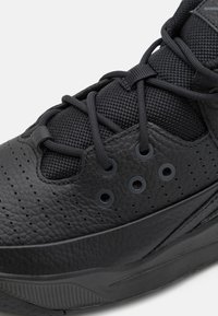 Gros plan d’une basket noire montrant du cuir texturé, des perforations, un tissu en maille et des lacets noirs passés dans des œillets ronds.