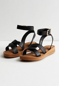 Mustat ristisangoilla varustetut sandaalit, joissa on ruskea pohja. Ominaisuuksiin kuuluu leveä risti-strap-muotoilu, nilkkahihna soljella ja rei'itetty pohja mukavuutta varten.