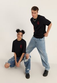 Zwei junge Erwachsene in schwarzen bedruckten T-Shirts und zerrissenen Jeans, einer hockt, der andere steht vor einem einfarbigen Hintergrund.