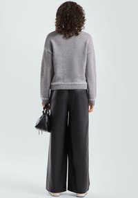 Grauer Strickpullover mit strukturierter Oberfläche, mit geripptem Saum und geometrischer Stickerei, kombiniert mit weiten schwarzen Hosen und einer kleinen Tasche.