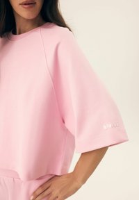 Różowa bluzka z krótkim rękawem uszyta z miękkiego materiału. Ma luźny krój, ribbowany dekolt oraz małe logo w subtelny ton na rękawie.