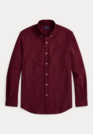 Jeune homme portant une chemise bordeaux, un pantalon plissé gris foncé, des chaussures marron et un pull vert porté sur les épaules, sur un fond uni.