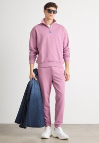Svetlo roza sweatshirt s polovičnim zadrgo in rebrastimi manšetami, v kombinaciji s ujemajočimi se trenirkami. Model drži jeans jakno in nosi bele superge.