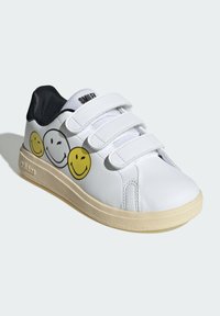 Bijele tenisice s tri Velcro trake, sa žutim grafičkim motivima nasmijanih lica, crnim akcentima i teksturiranom bež potplatom.