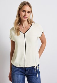 Gräddvit blus med korta ärmar, V-ringning, svarta pipings detaljer och dragsko i midjan. Bärs med blå denimjeans.