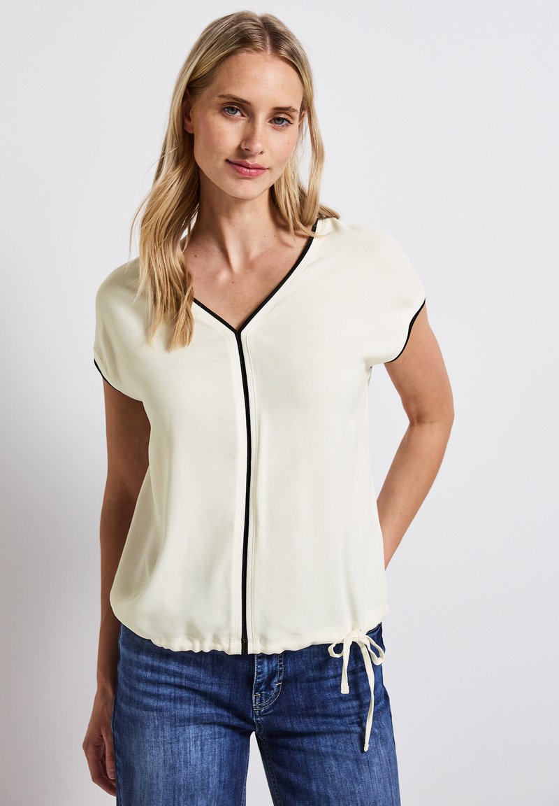 Gräddvit blus med korta ärmar, V-ringning, svarta pipings detaljer och dragsko i midjan. Bärs med blå denimjeans.