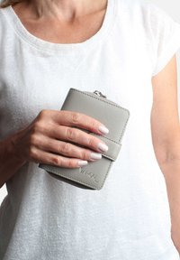 Gray leather wallet with a smooth texture, featuring a secure closure and embossed brand name. Held in a hand against a white shirt.