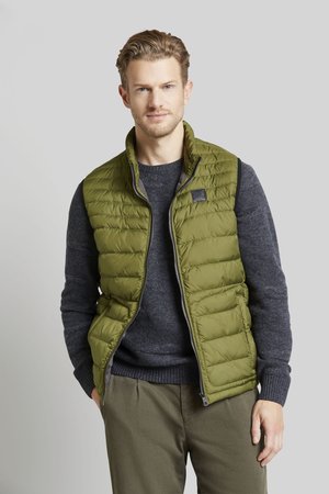 Olivgrüne Steppweste mit genähtem Muster, Reißverschluss vorne und Stehkragen, getragen über einem dunkelgrauen Pullover und kombiniert mit grüner Hose.